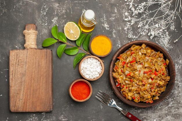 Top view plate of beans and spices bowls of colorful spices lemon the plate of green beans next to the cutting board bottle of oil and fork on the dark table