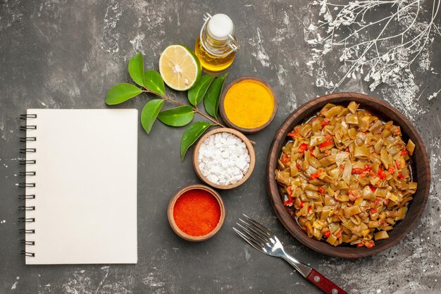 Top view plate of beans and spices bowls of colorful spices lemon white notebook the plate of green beans next to the bottle of oil and fork on the dark table