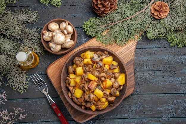 Вид сверху тарелка на борту тарелка с картофелем, грибами на деревянной доске рядом с вилкой под миской с грибным маслом в бутылке и ветвями деревьев с шишками