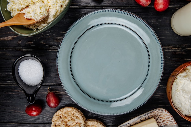 Top view of a plate , cottage cheese in a bowl, fresh sweet grapes and sugar in a saucer on dark wooden table