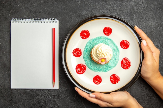 Piatto vista dall'alto del taccuino bianco cupcake e matita rossa accanto al cupcake sul piatto bianco nelle mani sullo sfondo scuro