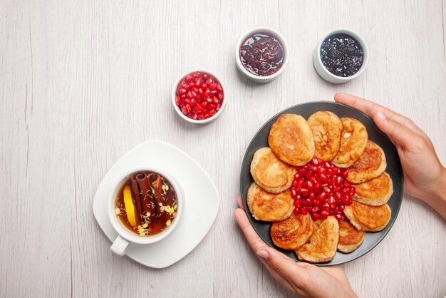 Top view plate in hands bowls of jam a cup of tea with lemon and plate of pancakes and pomegranate in hands on the table
