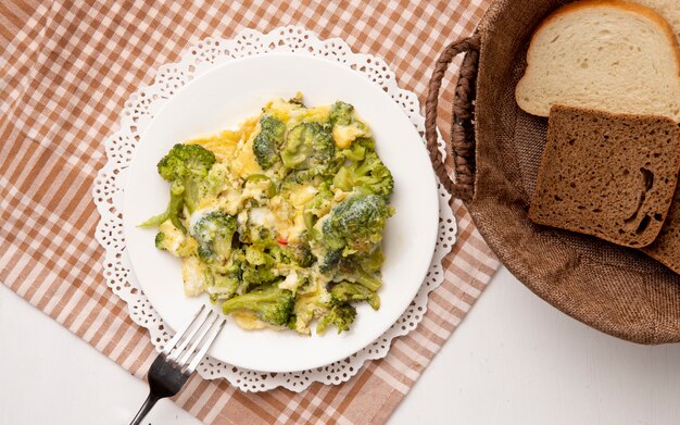 Top view of plate of meal with broccoli and eggs with fork on cloth and sliced breads on white background