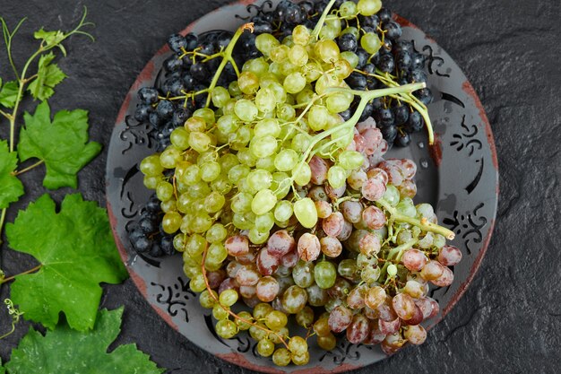Top view of a plate of mixed grapes around grape leaves on dark background. High quality photo