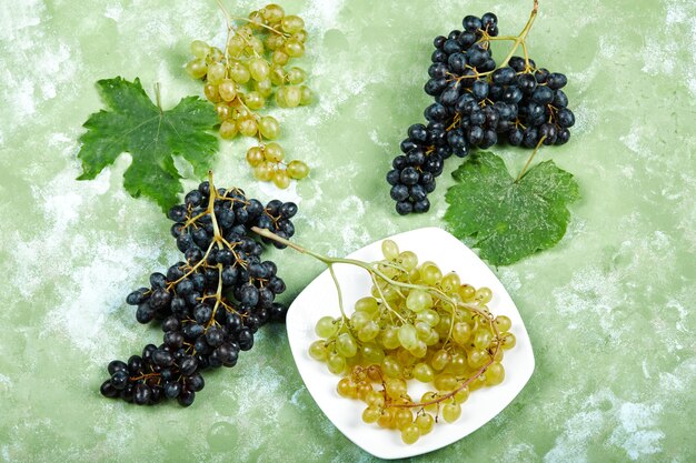 Top view of a plate of white grapes and black grapes with leaves on green surface