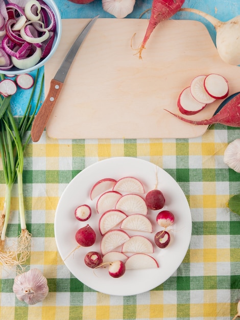 Top view of plate of whole and sliced radishes with scallions garlic and knife on cloth surface