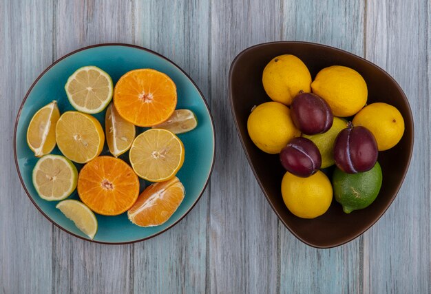 Вид сверху сливы в миске с дольками лимона, лаймом и апельсином на тарелке на сером фоне
