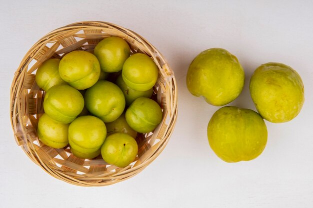 Top view of plums in basket and green pluots on white background