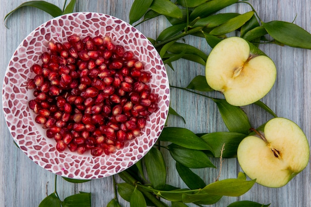 Top view of pomegranate berries in bowl and half cut apple with leaves on wooden surface