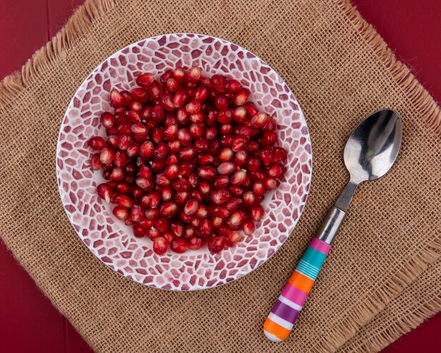 Top view of pomegranate berries in bowl with spoon on sackcloth on bordo surface