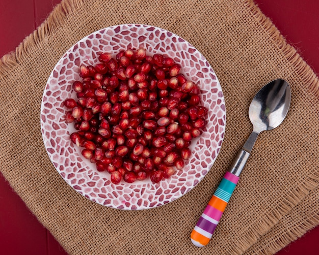 Top view of pomegranate berries in bowl with spoon on sackcloth on bordo surface