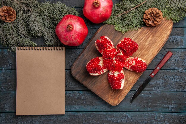 Top view pomegranate on board pilled pomegranate on cutting board next to spruce branches with cones knife and cream notebook on grey surface