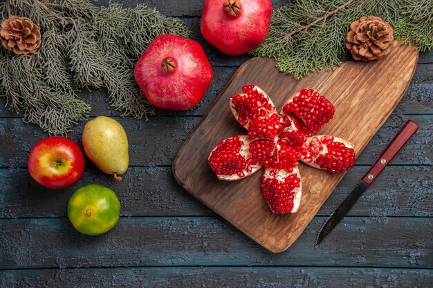 Top view pomegranate on board pilled pomegranate on cutting board next to spruce branches with cones knife lime apple pear on grey surface