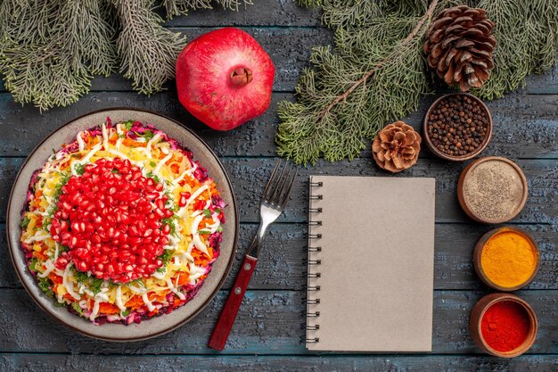 Top view pomegranate dish of potatoes seeds of pomegranate in the plate next to the fork white notebook pomegranate spruce branches with cones and colorful spices