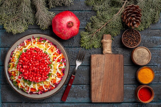 Top view pomegranate dish of potatoes seeds of pomegranate in the plate next to the fork wooden kitchen board pomegranate spruce branches with cones and colorful spices