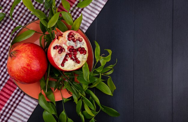 Free photo top view of pomegranate and half one with leaves in plate on plaid cloth on black surface