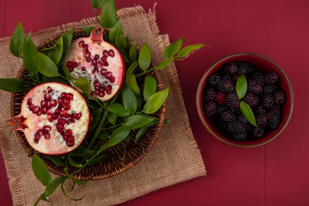 Top view of pomegranate halves with leaf branches in a basket of beige napkins with blackberries in a bowl on a red surface