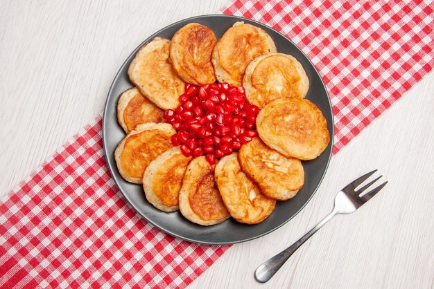 Top view pomegranate pancakes and seeds of pomegranate on the checkered tablecloth and a fork on the table