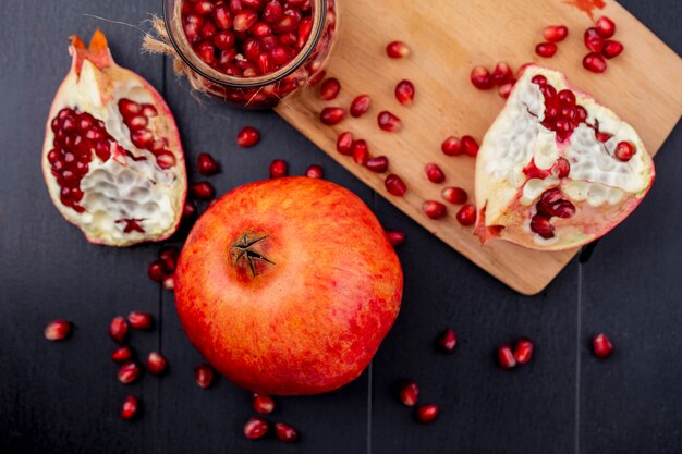 Top view of pomegranate piece and berries on cutting board and whole one with another piece on black surface