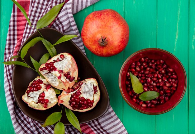 Top view of pomegranate pieces in bowl on plaid cloth with whole one and pomegranate berries in bowl on green surface