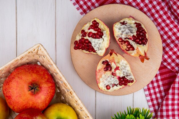 Top view of pomegranate pieces on cutting board on plaid cloth with whole one in basket on wooden surface