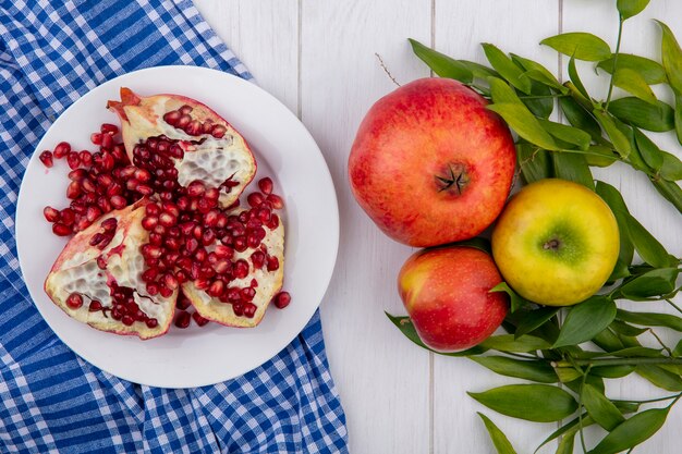 Top view of pomegranate pieces in plate on plaid cloth and whole one apple plum with leaves on wooden surface