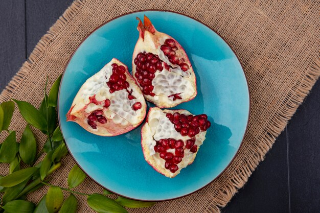 Top view of pomegranate pieces in plate with leaves on sackcloth and black surface