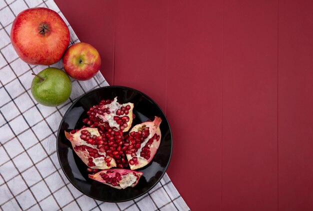 Top view of pomegranate pieces in plate with whole and apples on plaid cloth and bordo surface