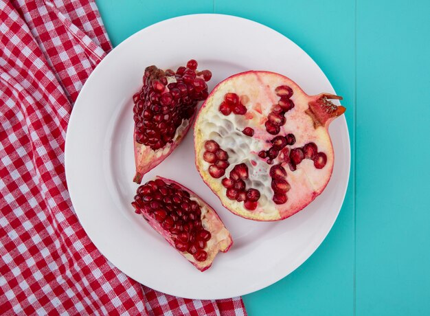 Top view of pomegranate pieces and pomegranate half in plate on plaid cloth on blue surface