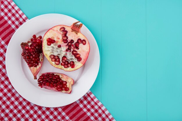 Top view of pomegranate pieces and pomegranate half in plate on plaid cloth on blue surface