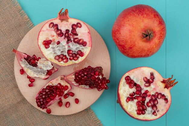 Top view of pomegranate pieces and pomegranate half with berries on cutting board on sackcloth with whole and half ones on blue surface