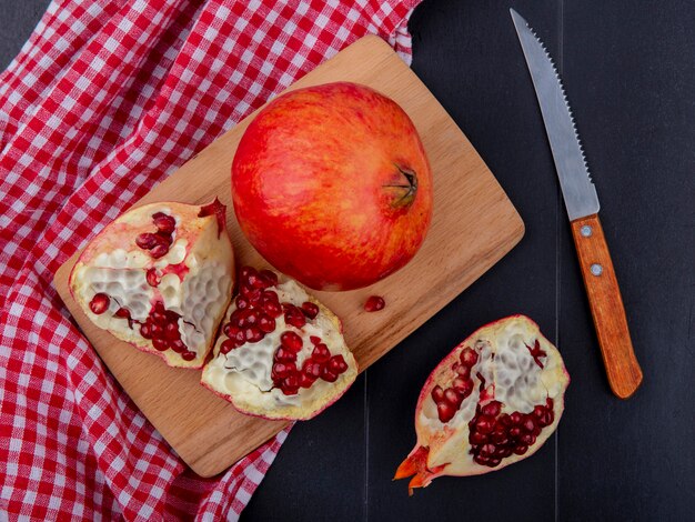 Top view of pomegranate pieces and whole one on cutting board on plaid cloth with knife on black surface