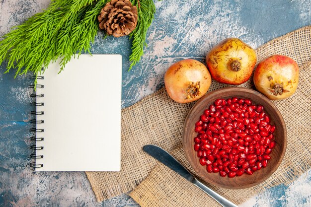 Top view pomegranate seeds in wooden bowl dinner knife pomegranates notebook pine tree branch on blue-white surface