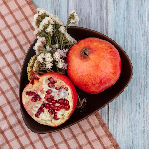 Top view of pomegranate with flowers in a bowl on a checkered brown towel on a gray surface