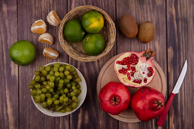 Top view pomegranates on a stand with green tangerines and grapes in a basket on a wooden table