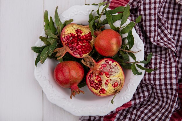 Top view pomegranates with leaves in a white plate and a checkered towel on a white background