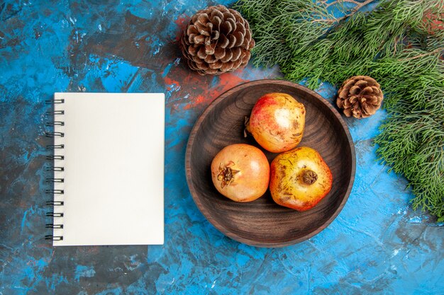 Top view pomegranates on wooden plate pine tree branch and cones a notebook on blue background