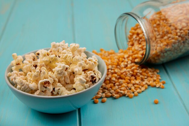 Top view of popcorns kernels falling out of a glass jar with popcorns on a bowl on a blue wooden table