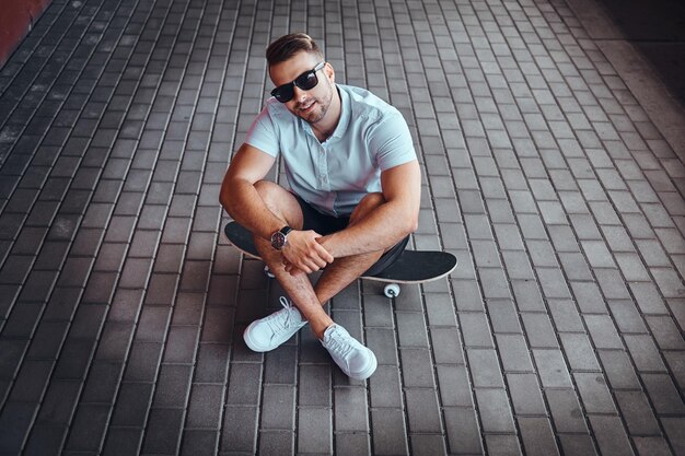 Top view portrait of a handsome fashionable skater guy in sunglasses dressed in a white shirt and shorts sitting on a skateboard on the footpath, looking at a camera.