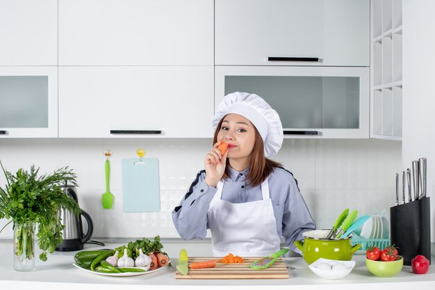 Top view of positive chef and fresh vegetables with cooking equipment and tasting carrot in the white kitchen