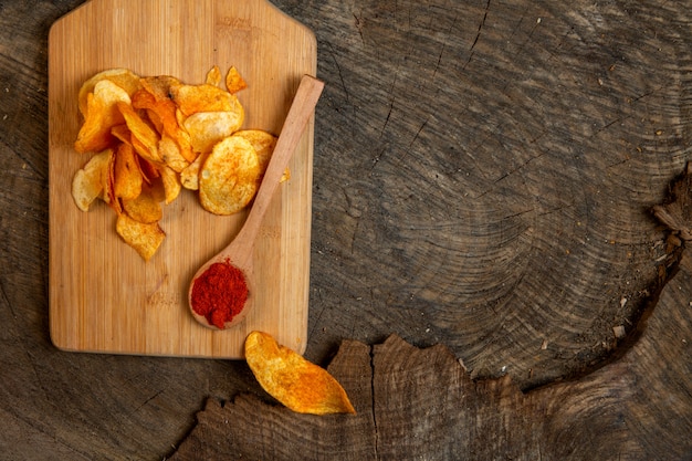 Top view of potato chips with a wooden spoon of chili pepper powder on a wooden cutting board with copy space