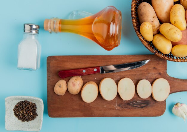 Top view of potato slices and knife on cutting board with whole ones in basket butter salt and black pepper and garlic on blue