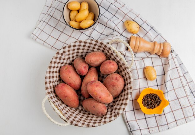 Top view of potatoes in basket and in bowl with salt black pepper on cloth on white surface