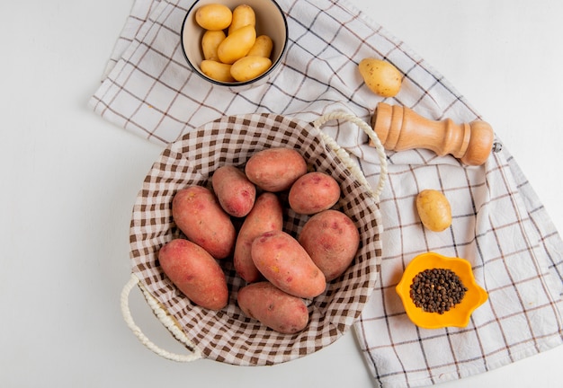 Top view of potatoes in basket and in bowl with salt black pepper on cloth on white