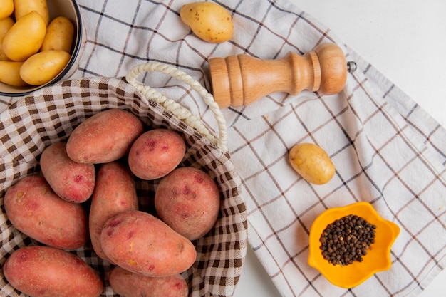 Vista superiore della merce nel carrello delle patate e in ciotola con pepe nero del sale sul panno del plaid su bianco