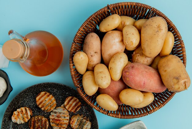 Вид сверху картофеля в корзине и жареные в сковороде с топленым маслом на синем
