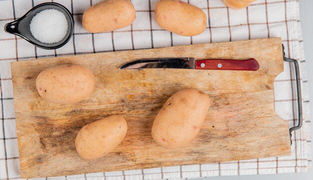 Top view of potatoes and knife on cutting board with other ones and salt on plaid cloth surface