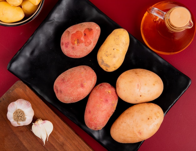 Top view of potatoes in plate with other ones in bowl melted butter and garlic on cutting board on bordo background