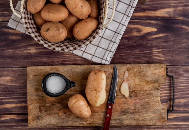 Top view of potatoes with shell knife and salt on cutting board with other ones in basket on cloth on wood