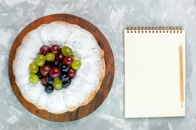 Вид сверху порошкообразный торт, вкусный запеченный торт со свежим виноградом и блокнотом на белом столе